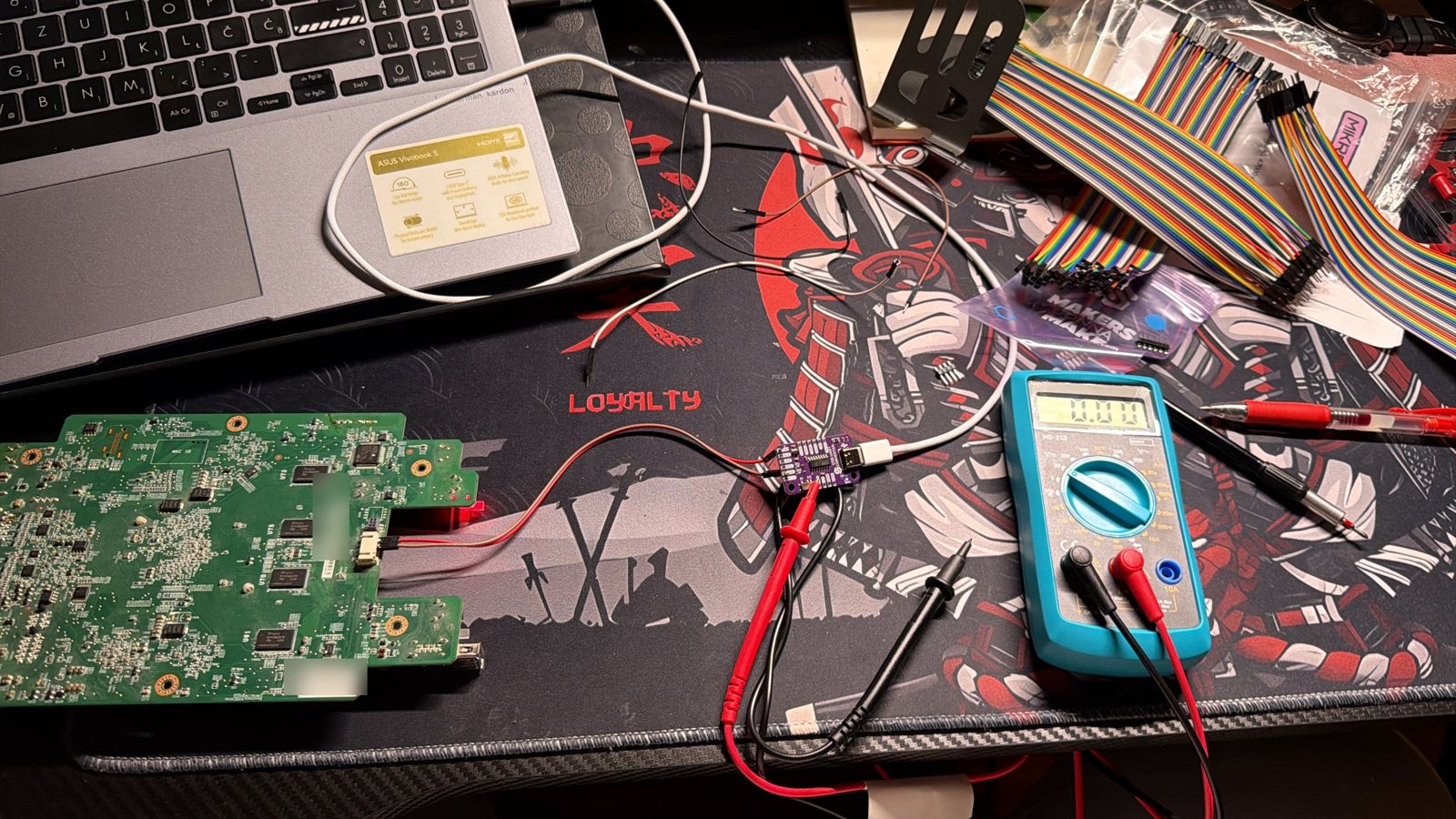 PR2100 motherboard connected by jumper wires to a small purple USB-to-UART adapter, with a multimeter next to it
